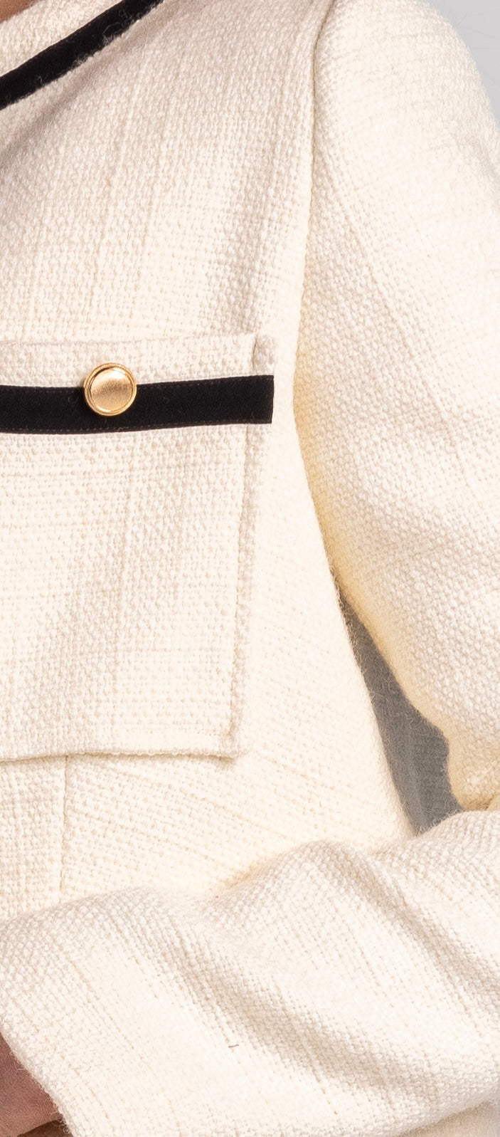 Close-up of a cream textured jacket with black trim and gold button on the chest pocket