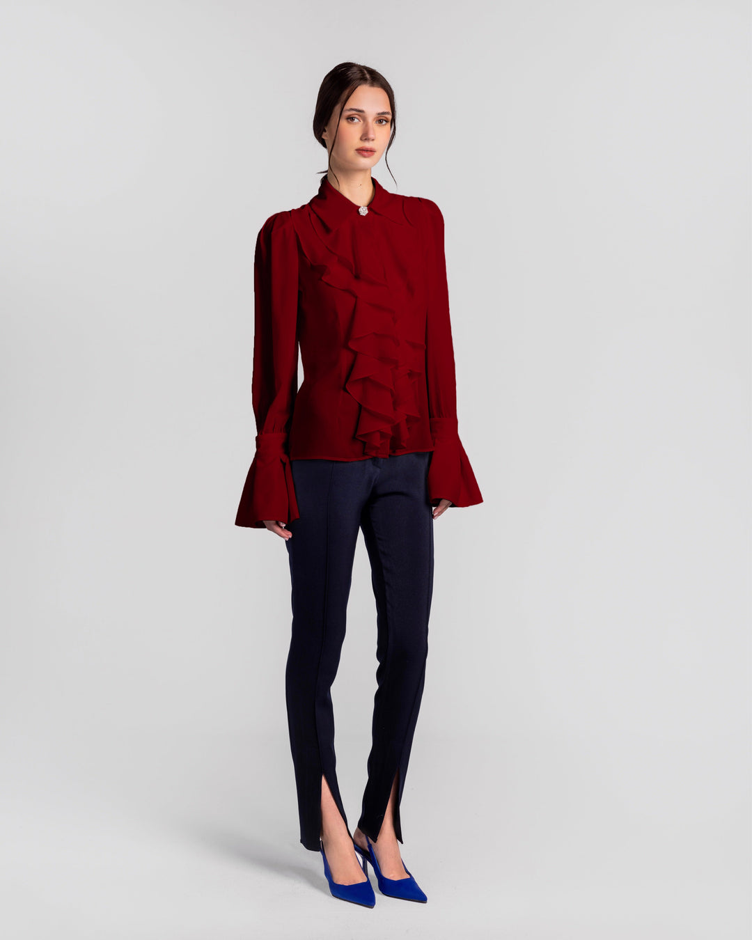 Woman in a red ruffle blouse, navy slim-fit split hem pants, and blue heels, studio background