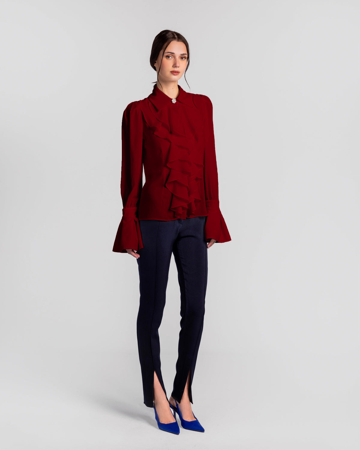 Woman in a red ruffle blouse, navy slim-fit split hem pants, and blue heels, studio background