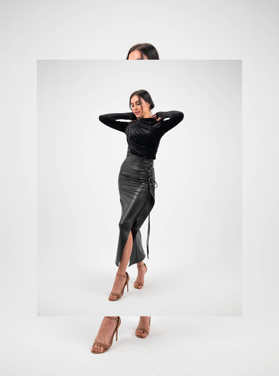 Woman in black velvet top and high-waisted faux leather midi skirt with side slit, neutral heels, studio background