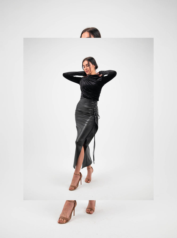 Woman in black velvet top and high-waisted faux leather midi skirt with side slit, neutral heels, studio background