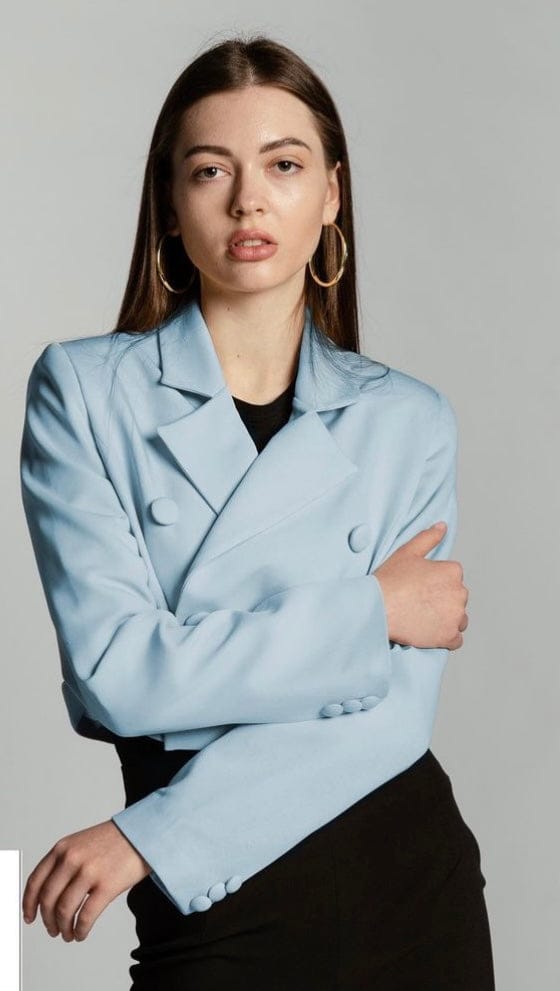 Woman wearing a light blue cropped blazer and black top, posing against a gray background