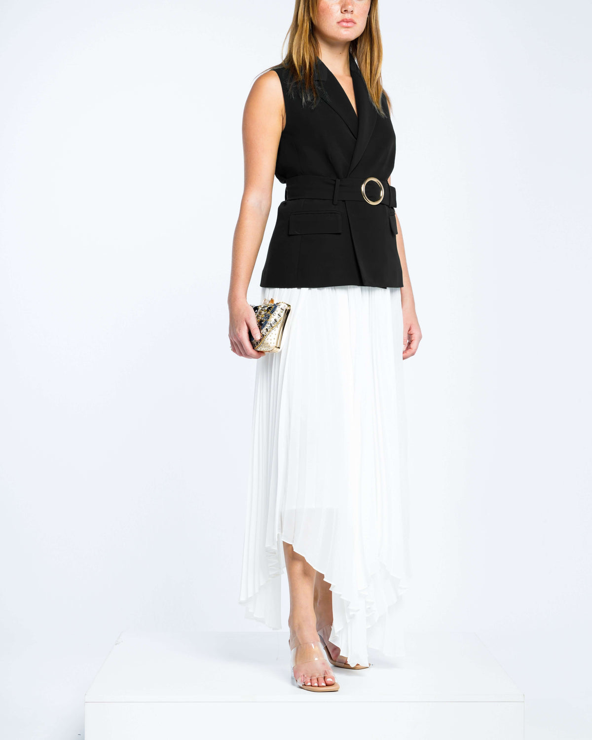 Woman wearing sleeveless black belted blazer and white pleated midi skirt, holding clutch bag