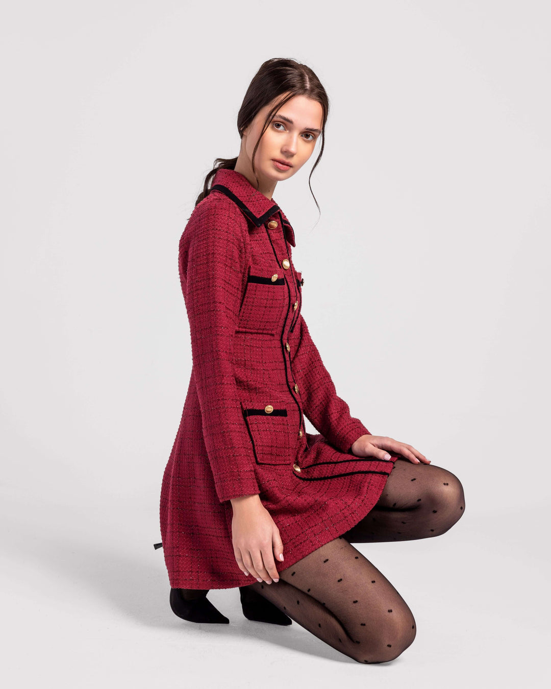 Woman in a red tweed dress with black trim, black polka dot tights, kneeling on plain background