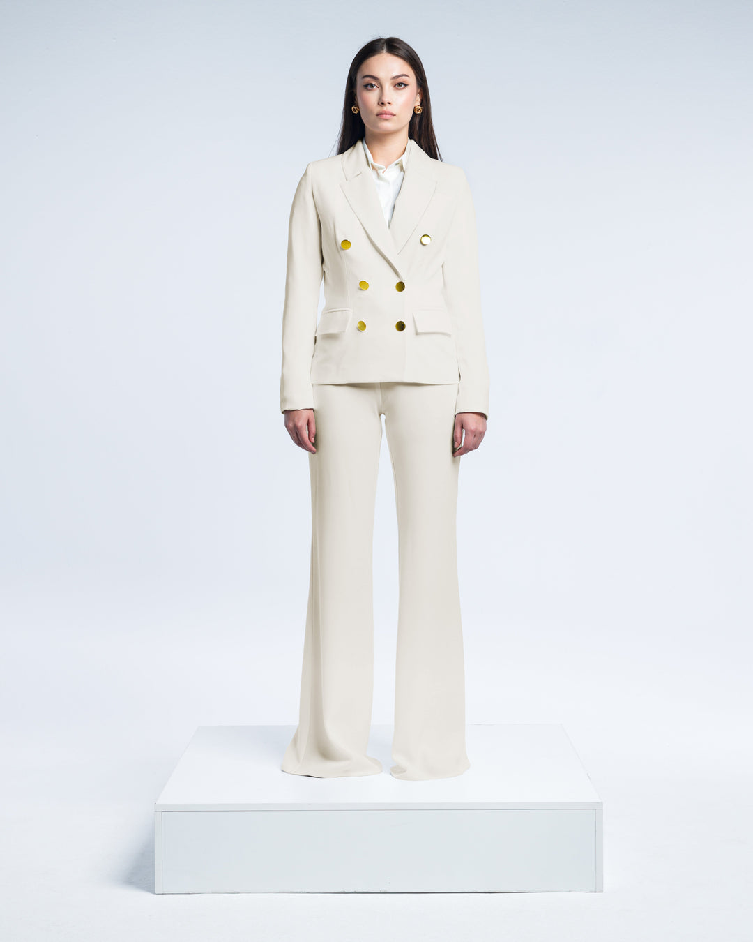 Woman wearing elegant beige pantsuit with gold buttons, standing in a white studio