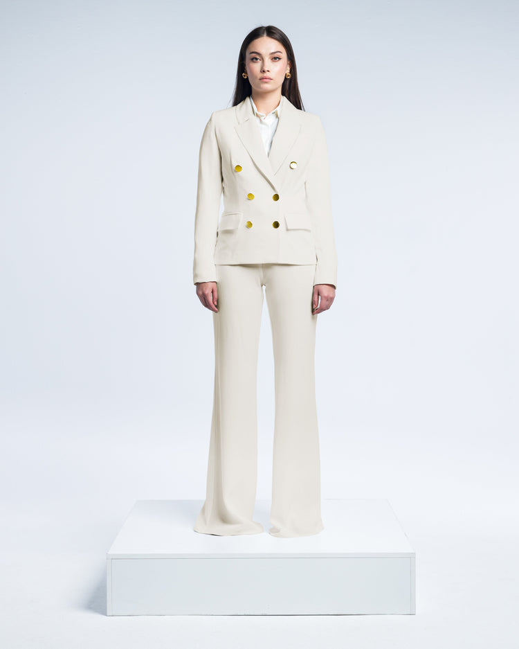 Woman wearing elegant beige pantsuit with gold buttons, standing in a white studio