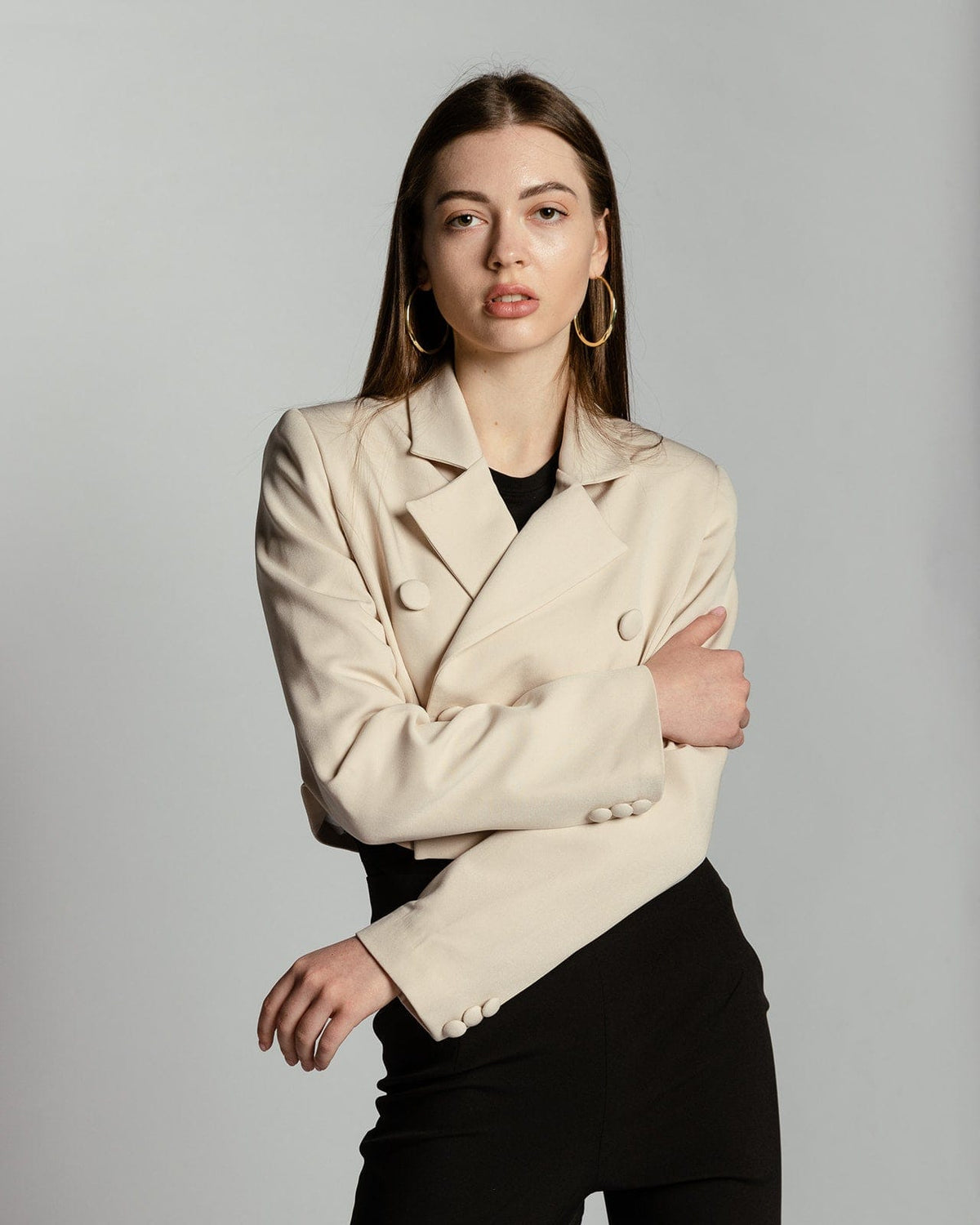 Woman wearing a beige double-breasted blazer with black pants, gold hoop earrings, and neutral makeup against a plain background.