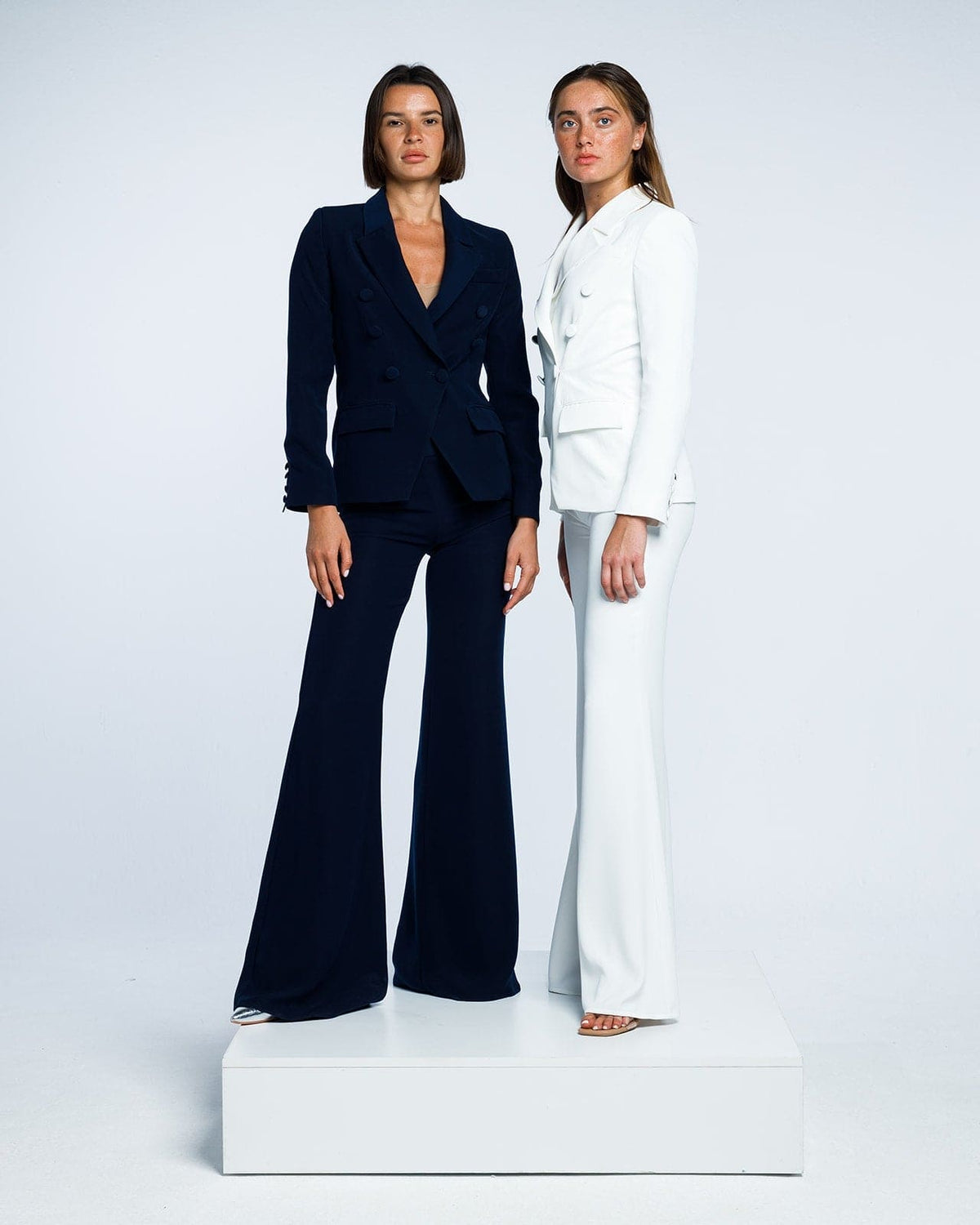 Two women wearing tailored double-breasted suits in black and white, posing on white platform