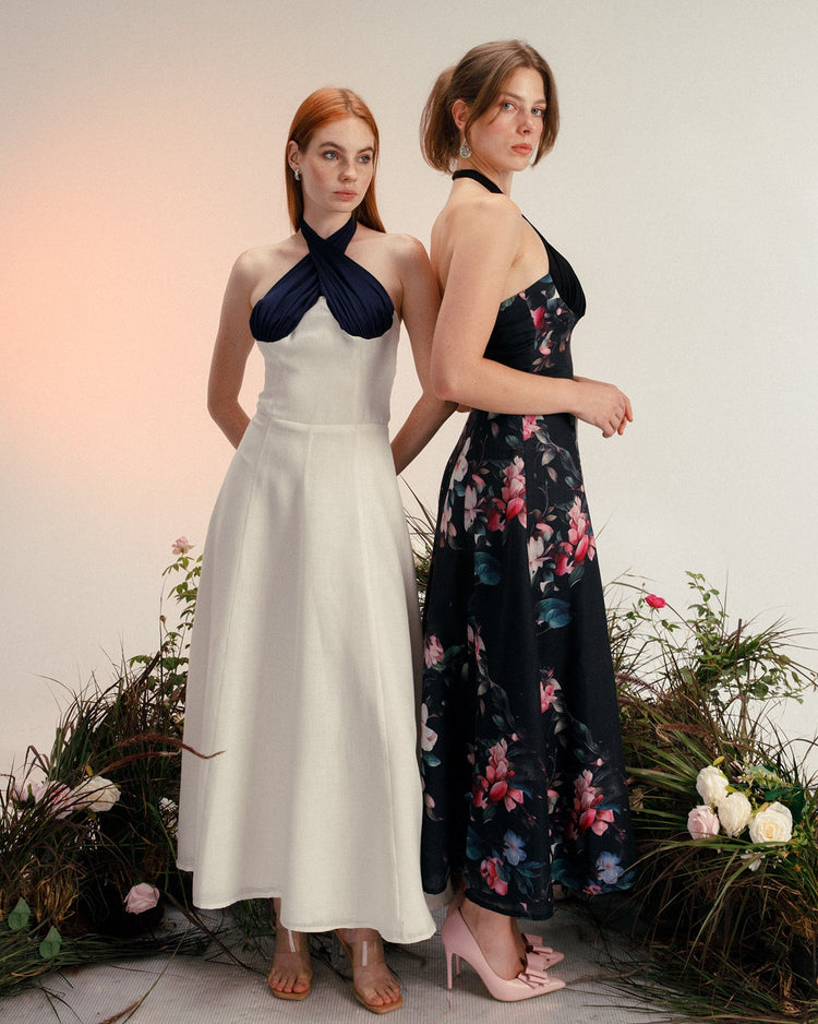 Two women modeling elegant halter neck dresses, one in white and navy, one in black floral, standing among flowers.