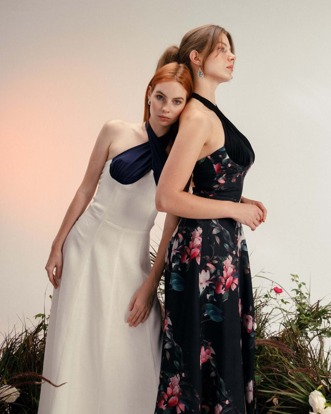 Two women in elegant halter neck dresses, one in white and one in black floral, posing indoors