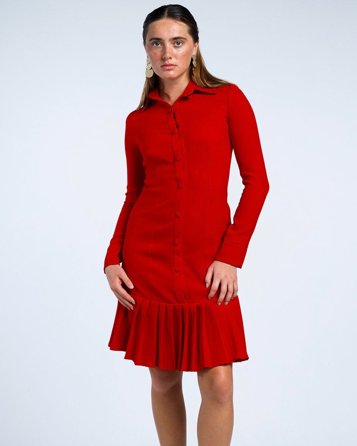 Woman in a fitted red long-sleeve dress with pleated hem, standing against white background.