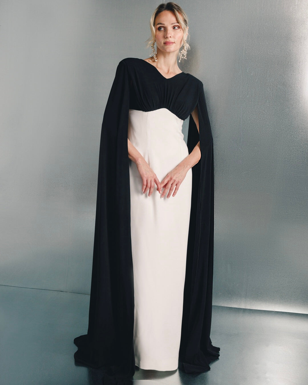 Woman in an elegant black and white cape evening gown with statement earrings, standing against a metallic background.