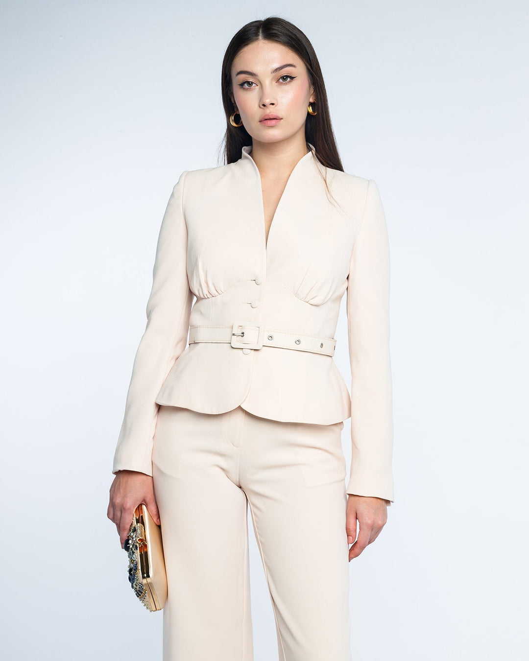 Woman wearing a beige tailored blazer and pants set with a belted waist and holding a jeweled clutch