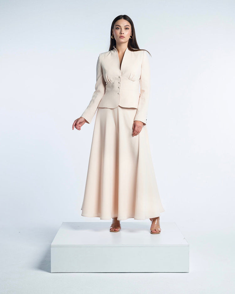 Woman modeling elegant beige suit with long-sleeve jacket and flowing maxi skirt on white platform