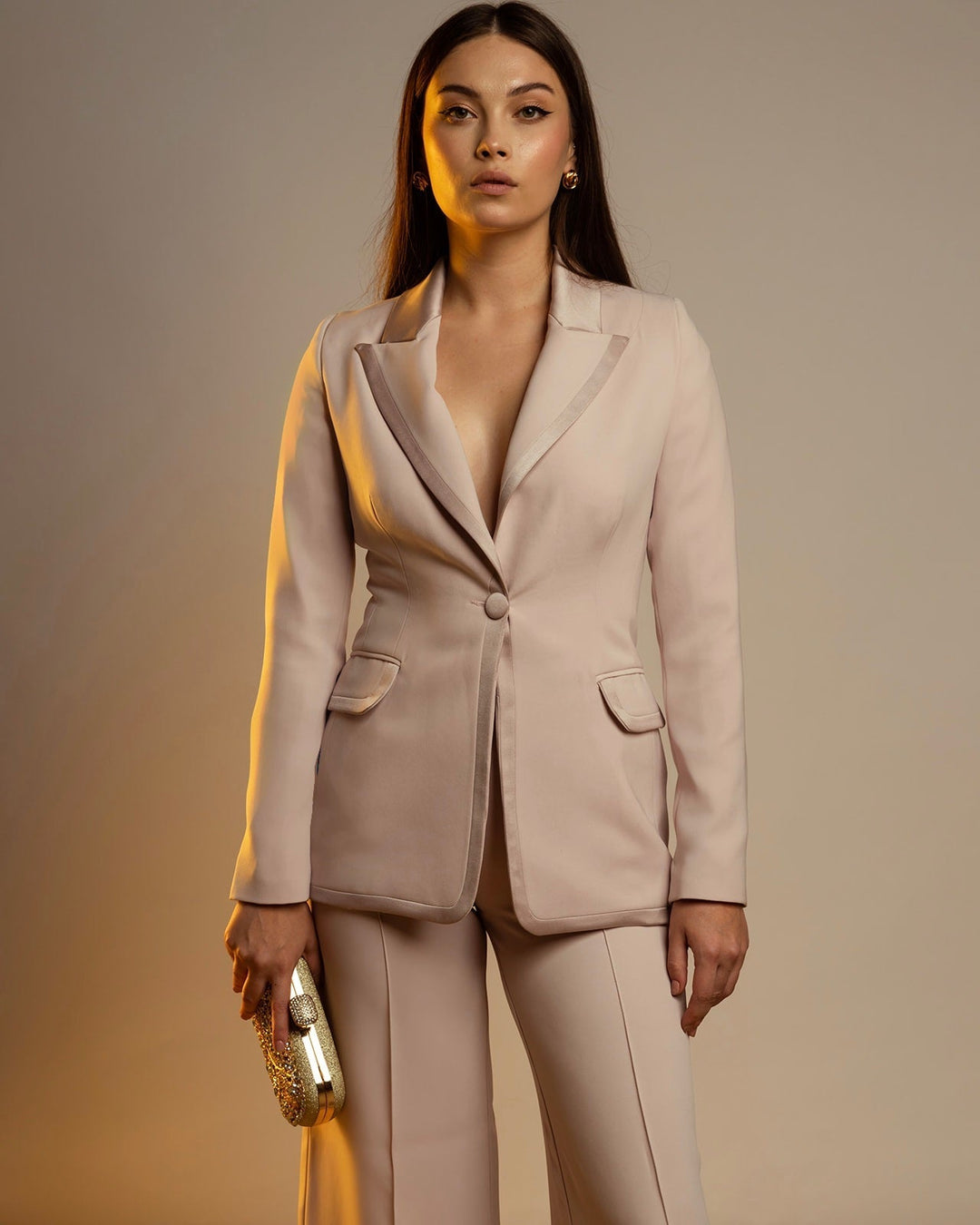 Woman in a cream suit with wide-leg pants holding a gold clutch in a studio setting