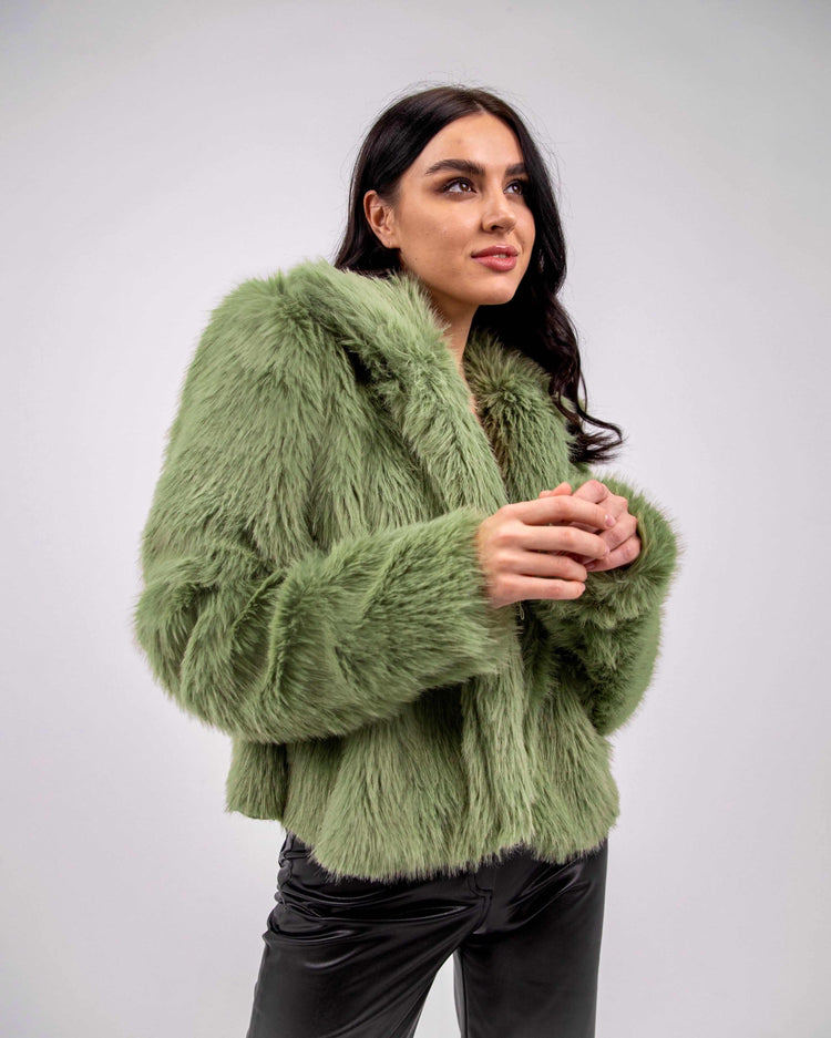 Woman wearing an olive green faux fur jacket and black pants against a plain background
