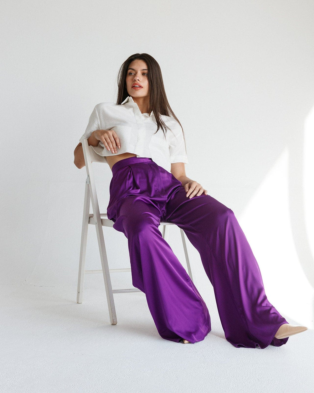 Woman in white cropped blouse and wide-leg purple satin pants sitting on chair in minimal studio