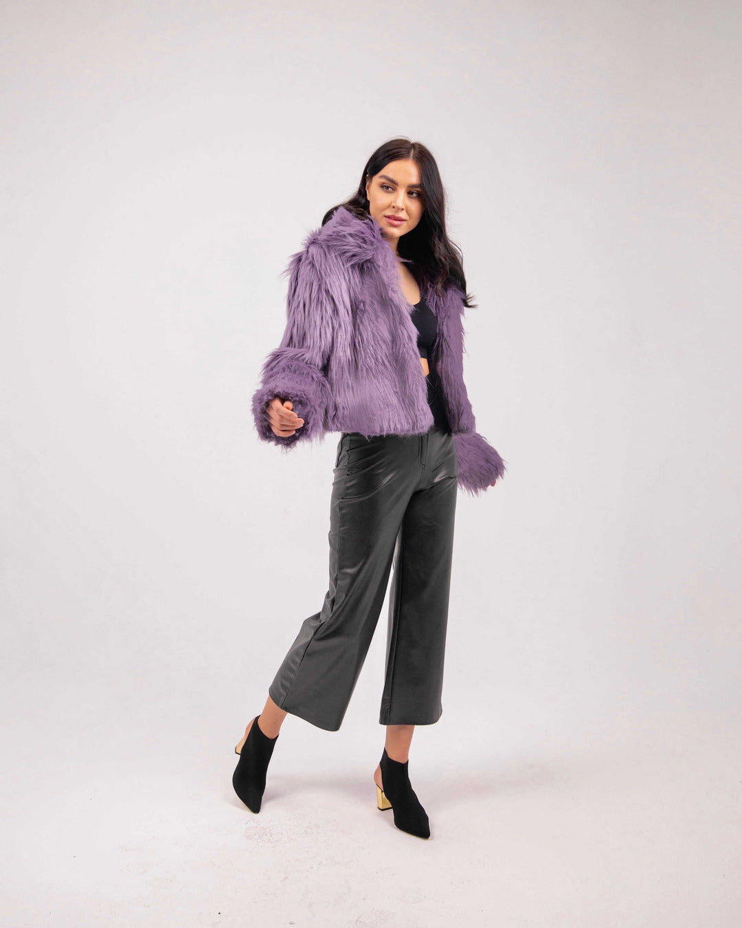 Woman wearing a purple faux fur jacket, black crop top, leather pants, and ankle boots