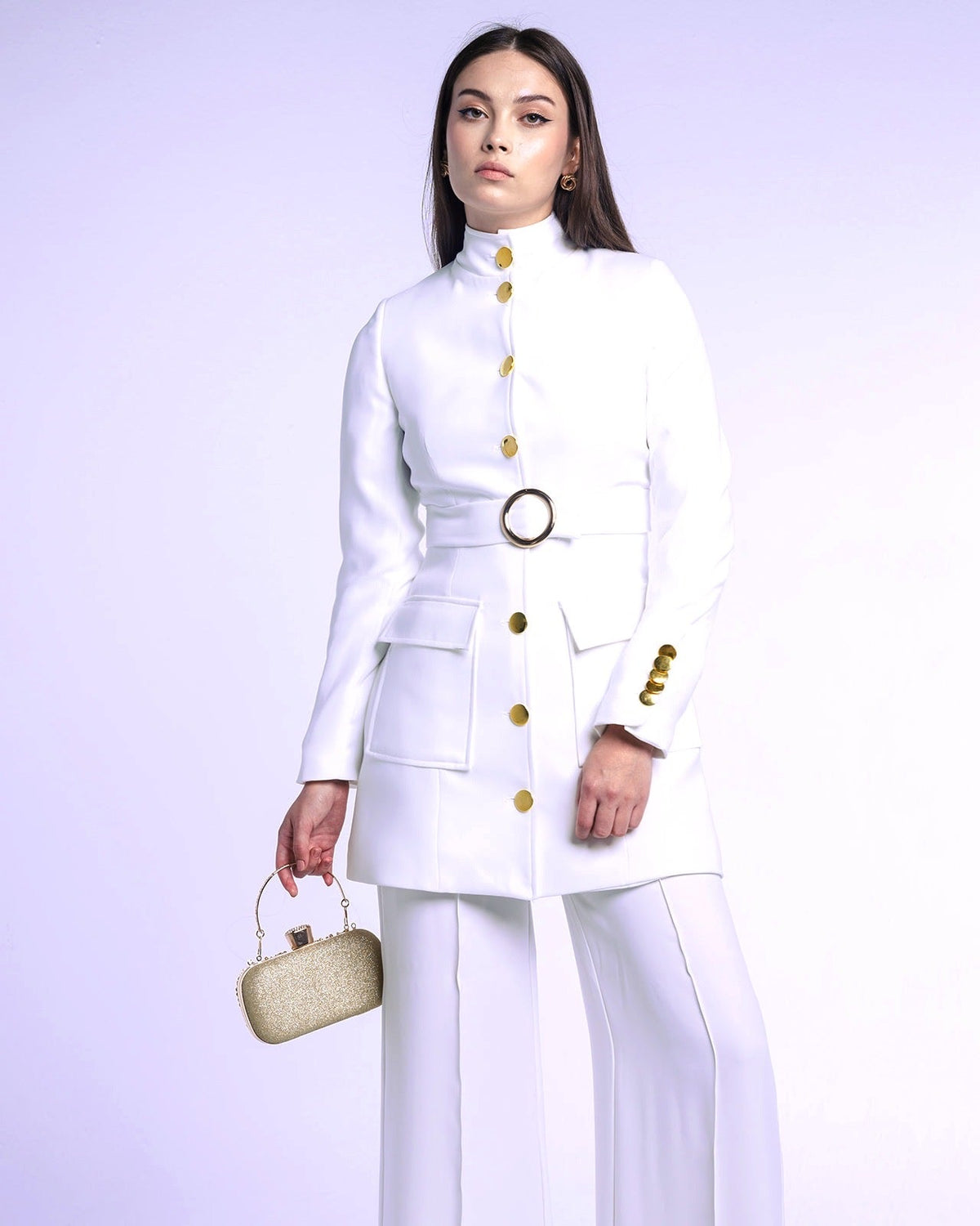Woman in white tailored suit with gold buttons and belt, holding gold clutch bag, fashion studio photo.