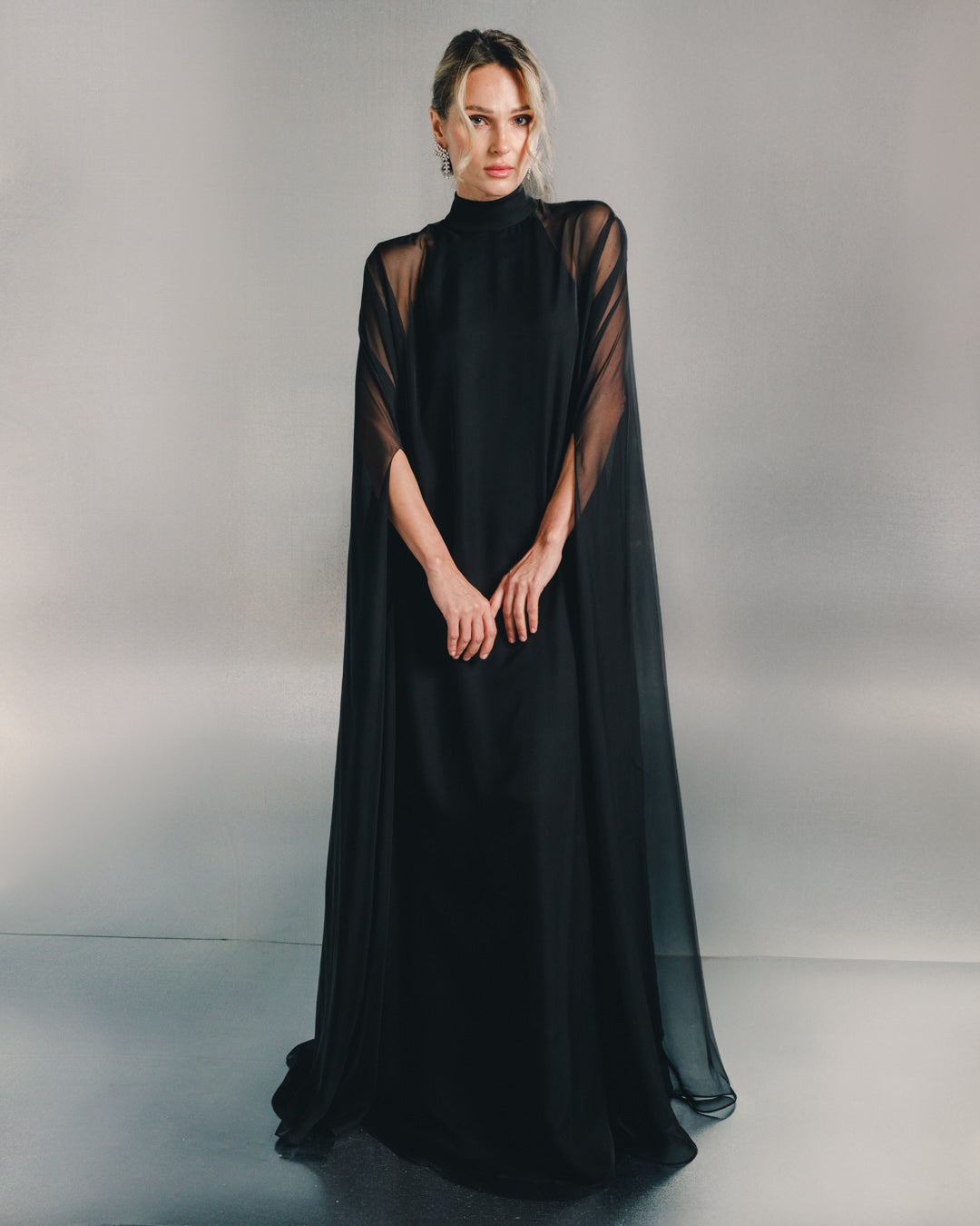 Woman in elegant black sheer cape dress with high neckline, standing against neutral background