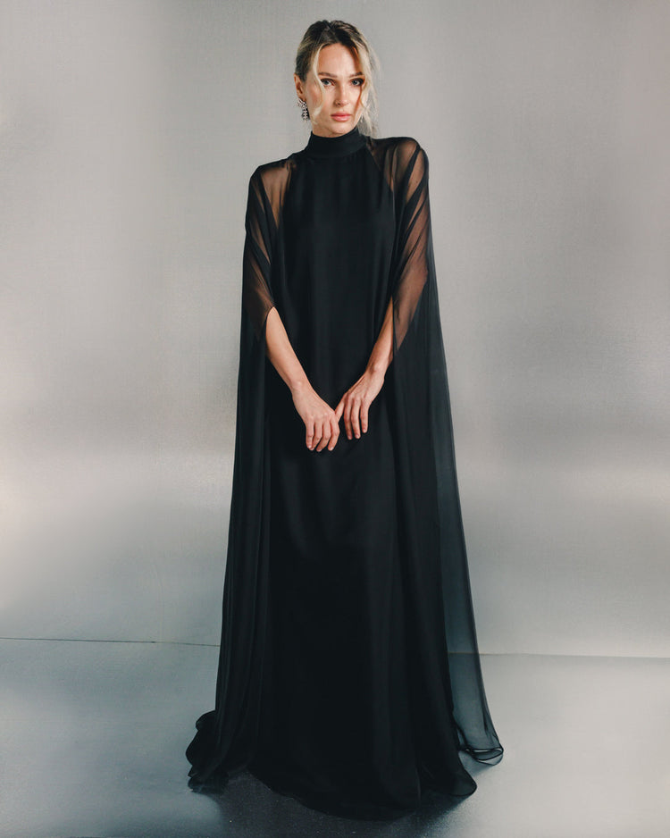 Woman in elegant black sheer cape dress with high neckline, standing against neutral background