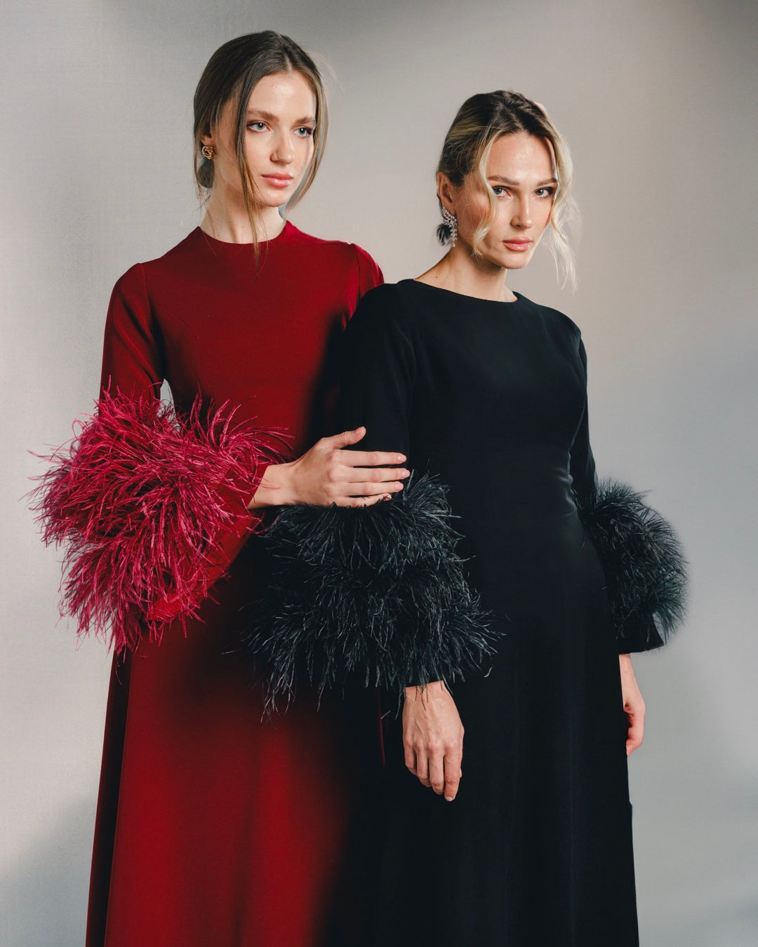 Two women in elegant long-sleeve gowns with feathered cuffs, one in red, one in black.