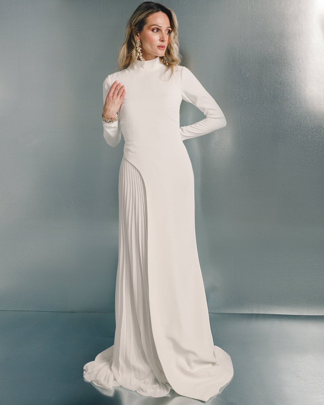 Woman wearing elegant long white gown with high neck and pleated skirt panel, metallic backdrop
