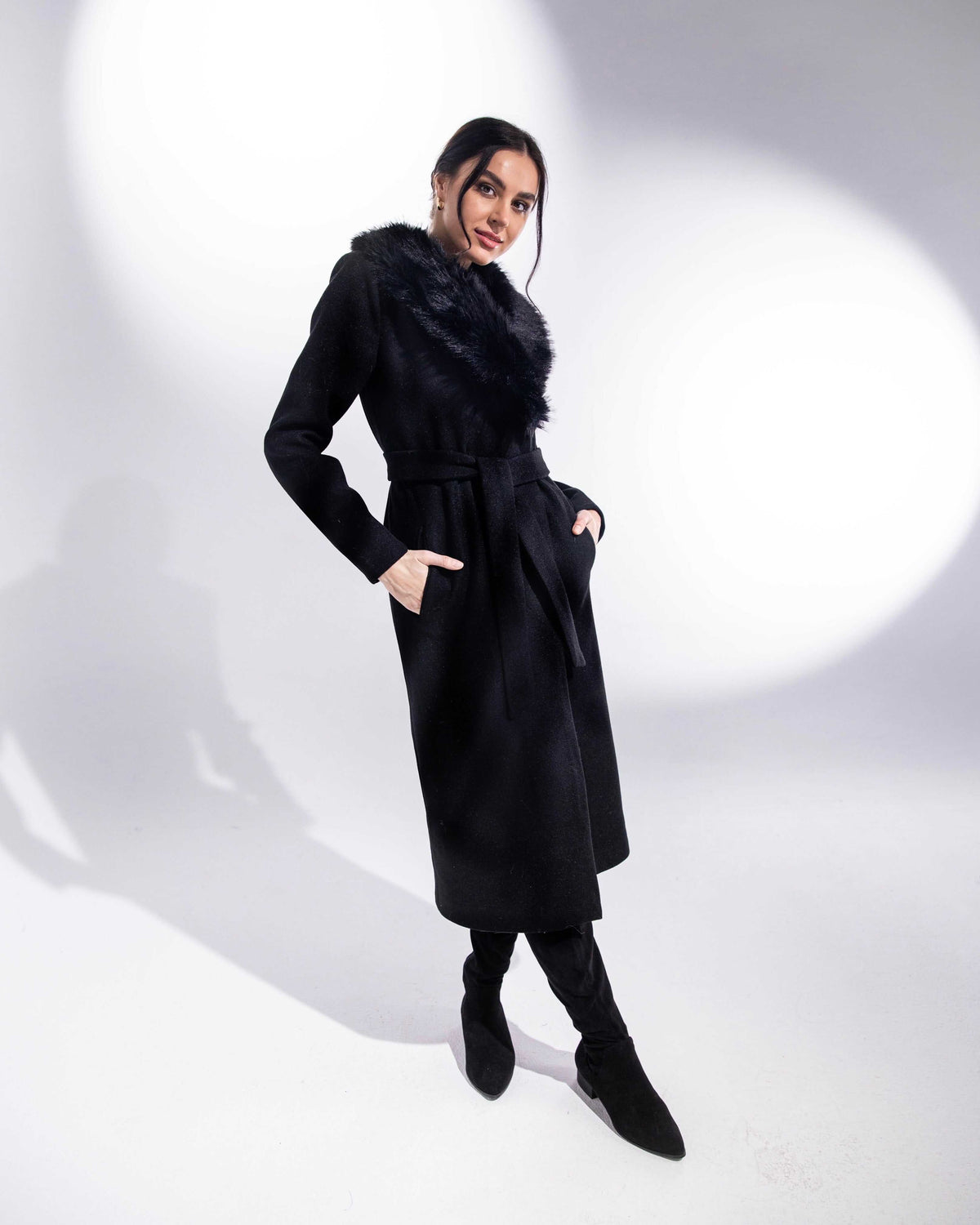 Woman modeling elegant black long coat with faux fur collar and belted waist in studio