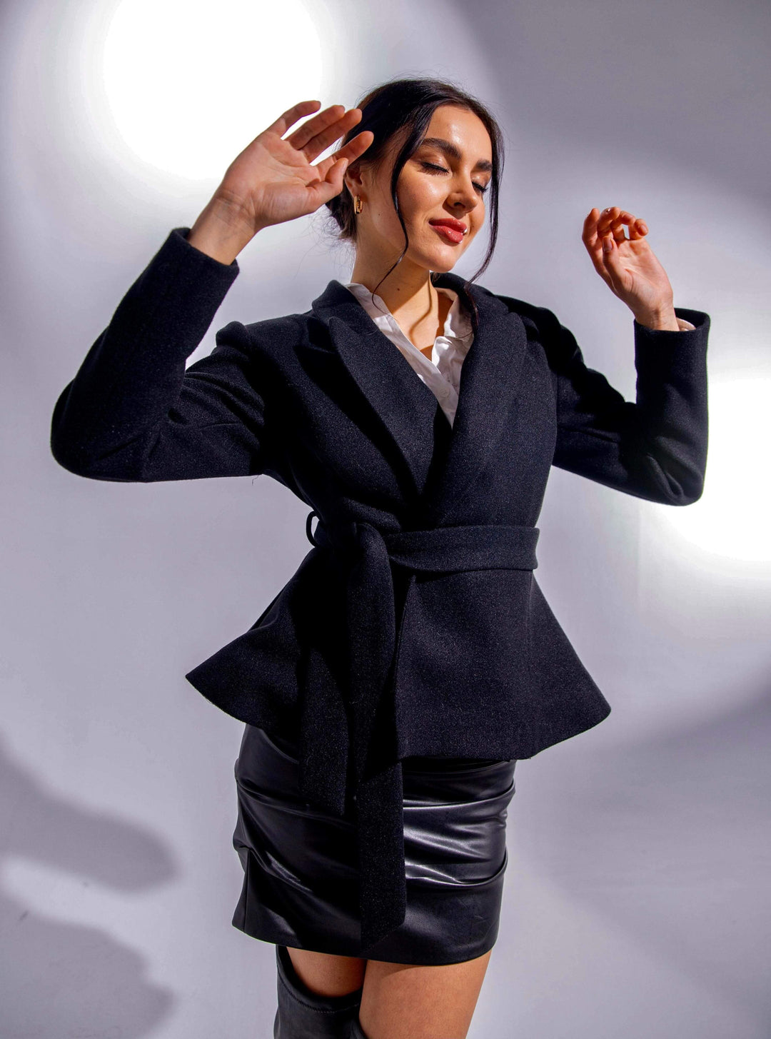 Woman modeling black belted blazer with faux leather mini skirt in studio lighting