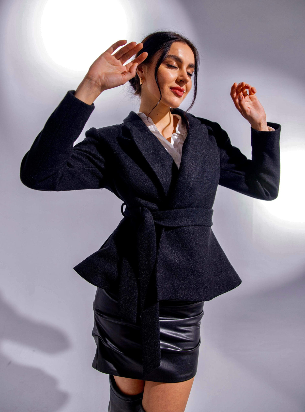 Woman modeling black belted blazer with faux leather mini skirt in studio lighting