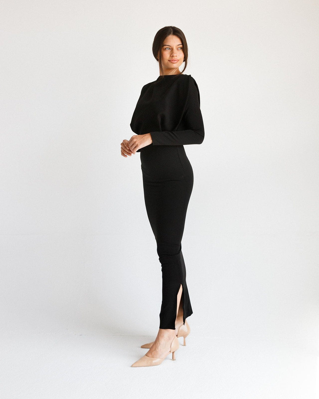 Woman wearing a long black fitted dress with long sleeves and side slit, standing in a minimal studio