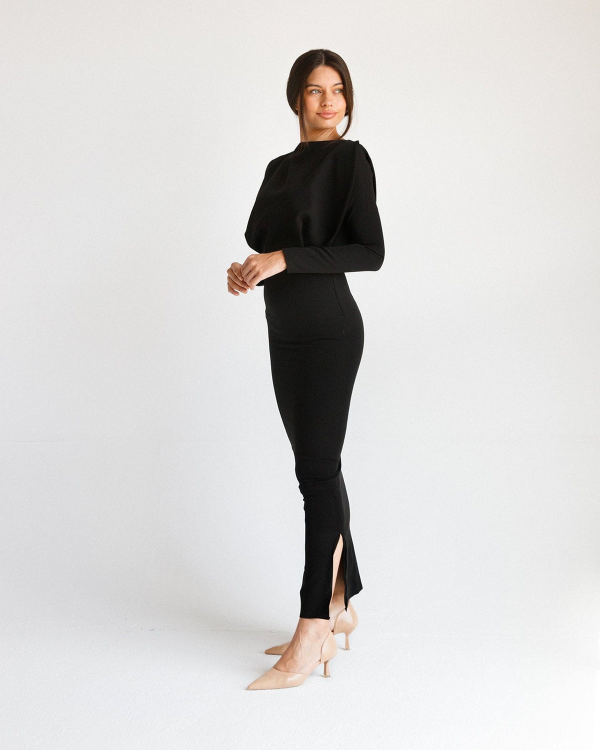 Woman wearing a long black fitted dress with long sleeves and side slit, standing in a minimal studio