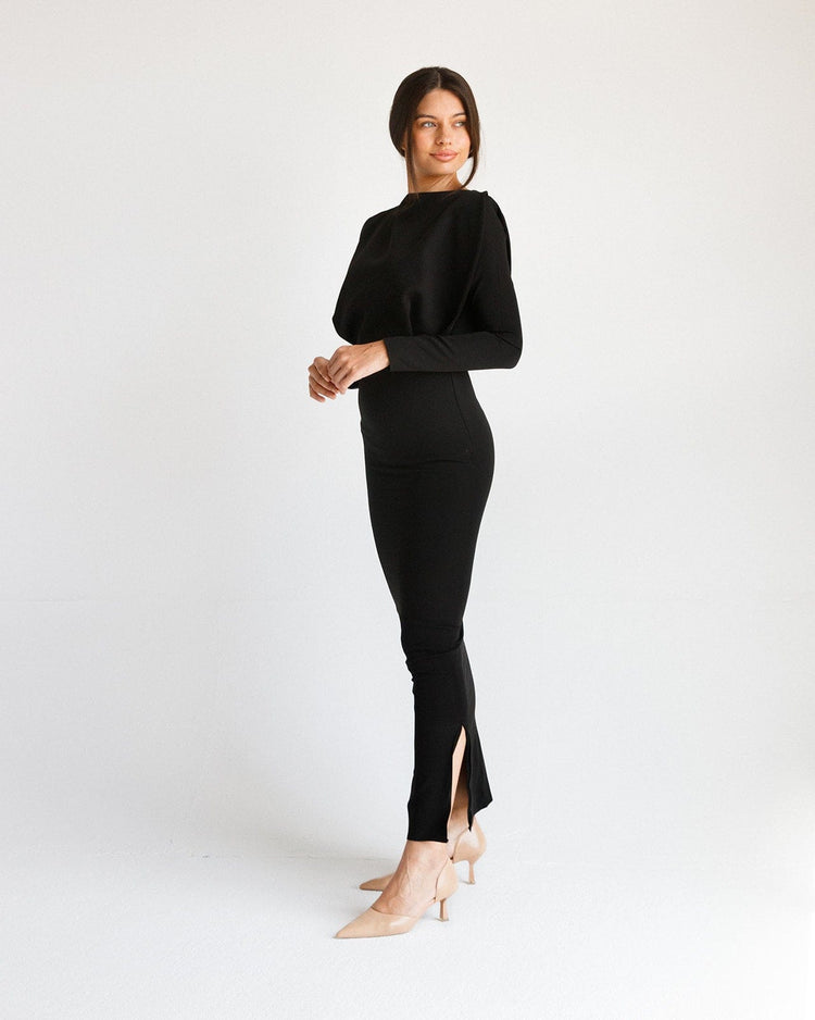 Woman wearing a long black fitted dress with long sleeves and side slit, standing in a minimal studio