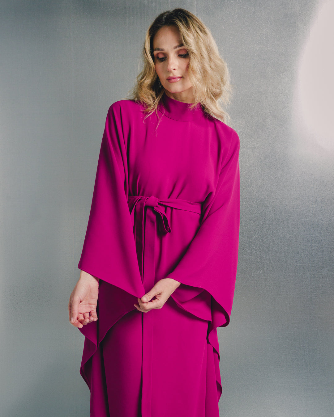 Woman wearing a fuchsia long-sleeve belted dress with draped sleeves against a silver background