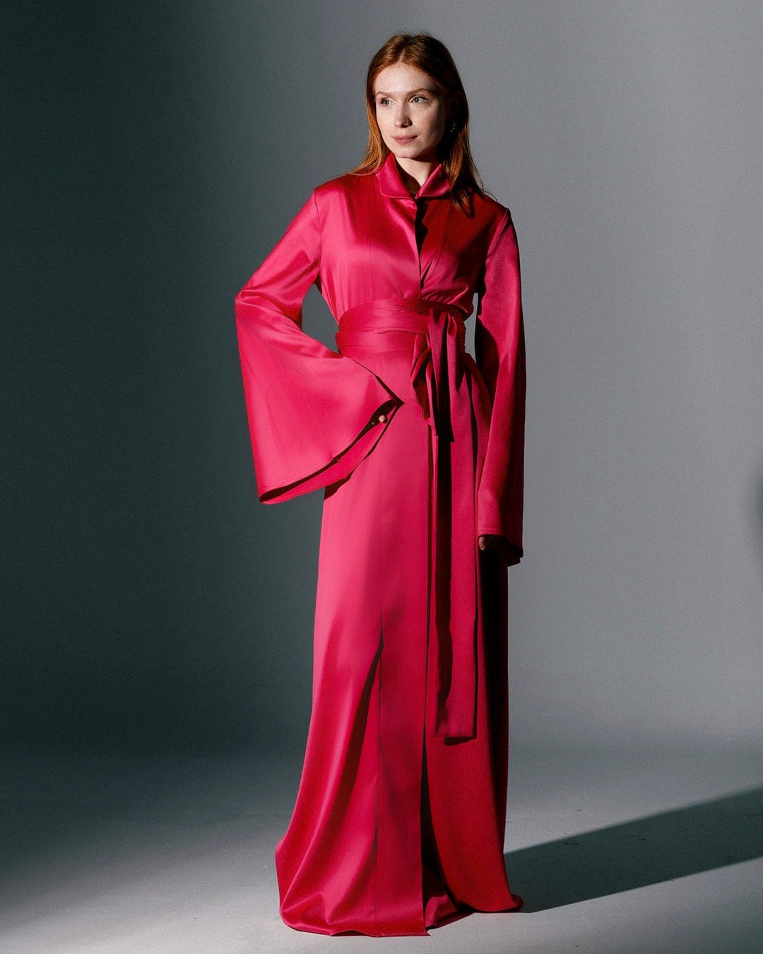 Woman wearing a fuchsia long satin kimono with wide sleeves and waist tie in studio setting