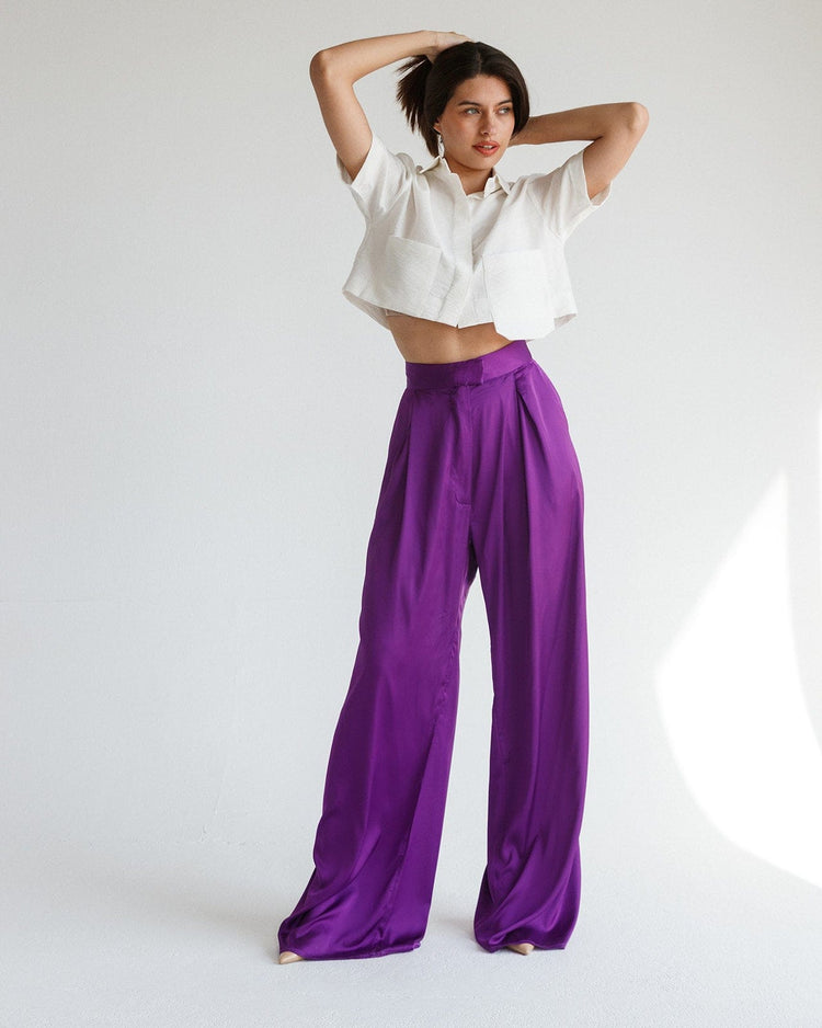 Woman in white cropped shirt and high-waisted purple satin wide-leg pants, standing indoors.