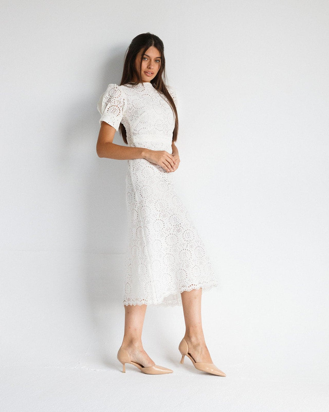 Woman wearing elegant white lace midi dress with puff sleeves and nude heels, standing against white background