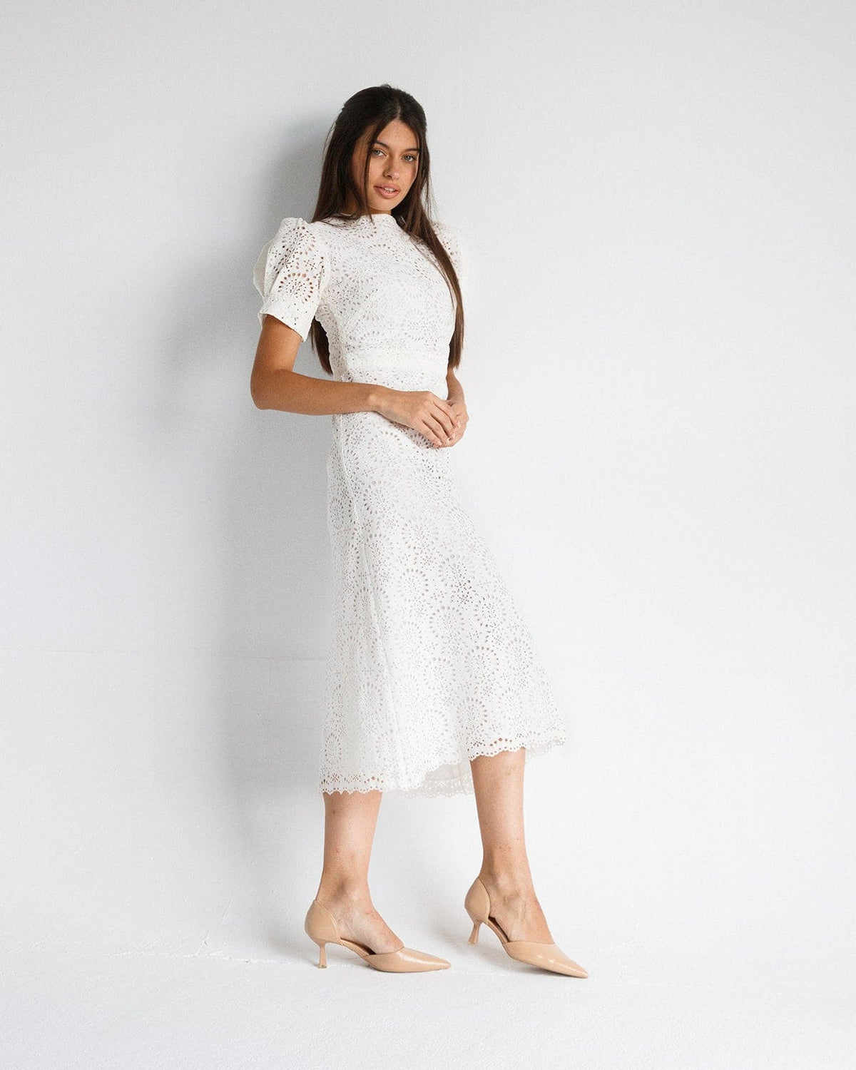Woman wearing elegant white lace midi dress with puff sleeves and nude heels, standing against white background