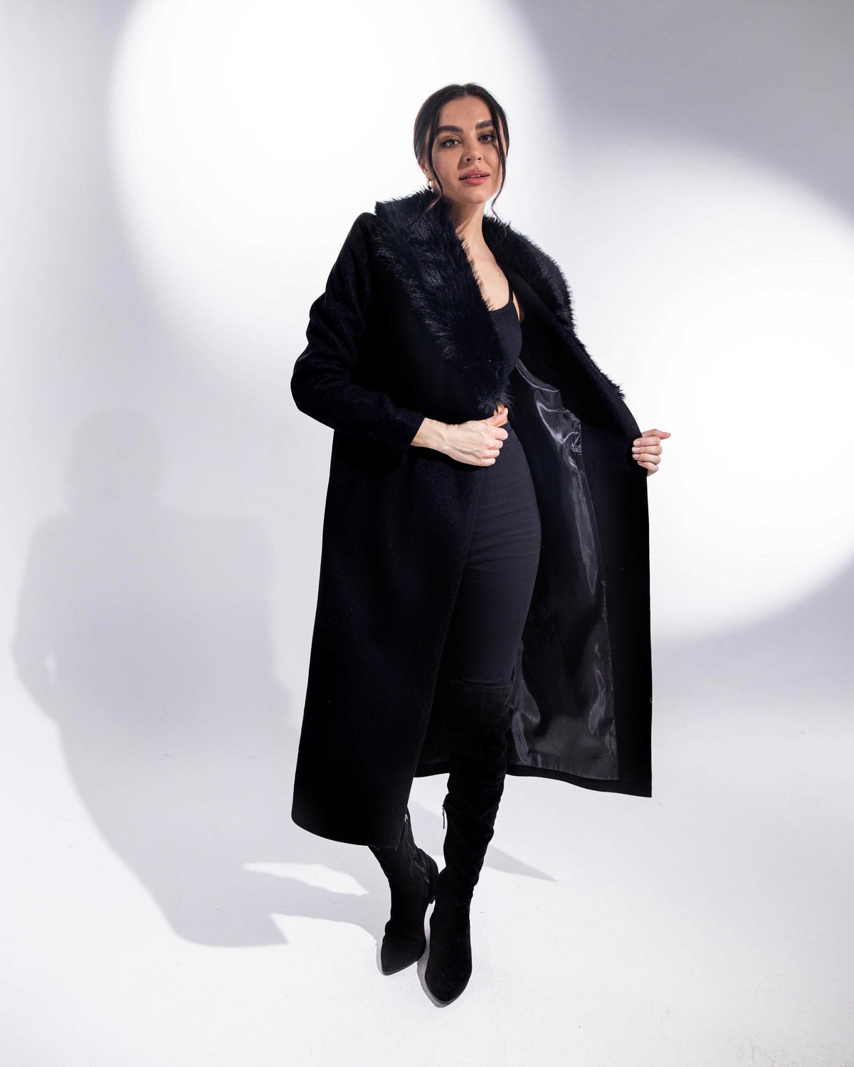 Woman modeling elegant black faux fur collar coat with thigh-high boots in studio