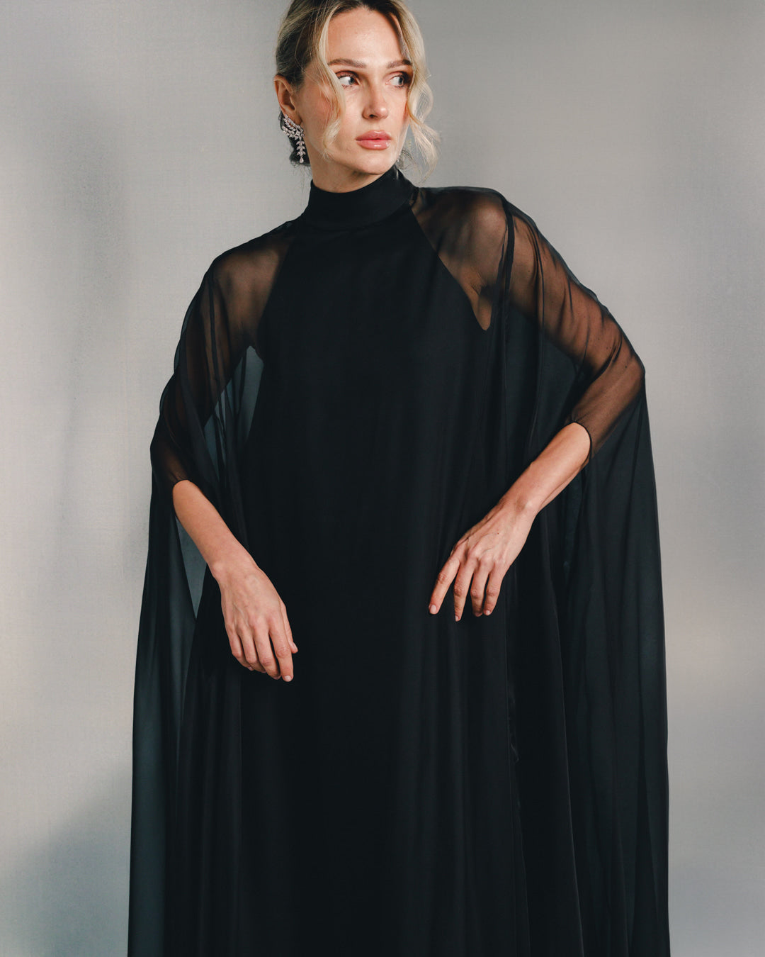 Woman in elegant black sheer cape dress with high neck and statement earrings