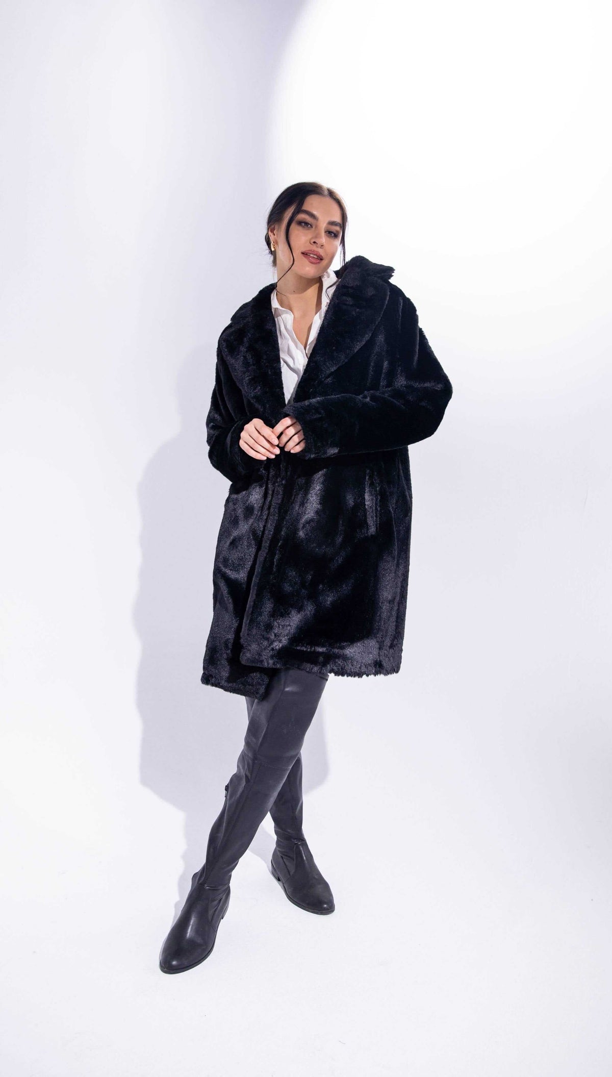 Woman wearing a black faux fur coat, white blouse, and black knee-high boots on white background
