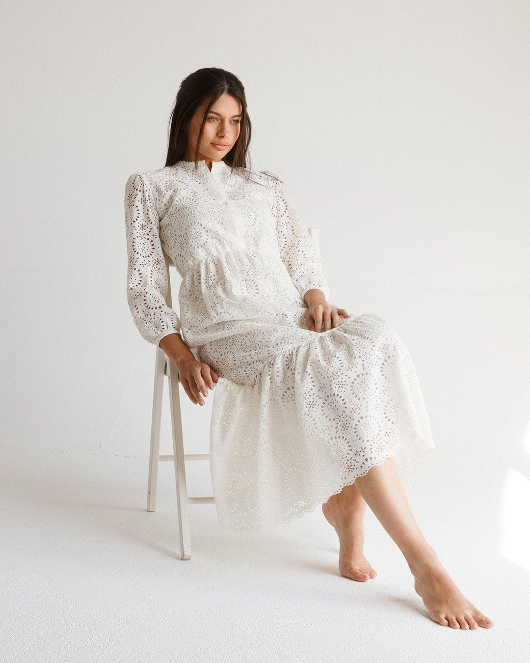 Woman in a long white lace dress with puff sleeves sitting barefoot on a chair in a bright studio