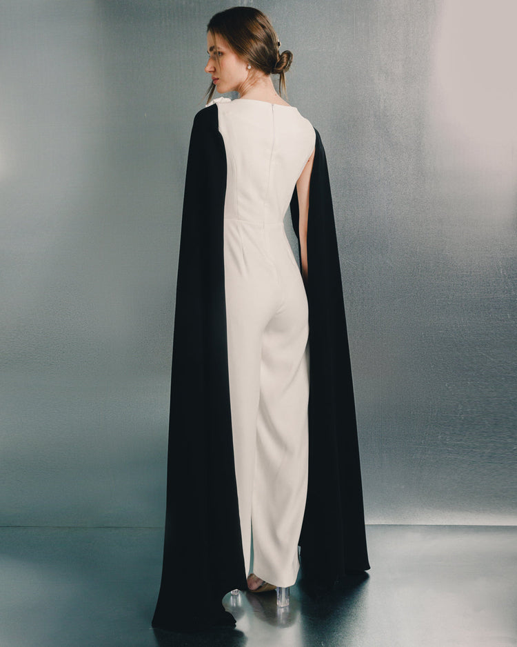 Woman in a white jumpsuit with black cape, standing in a modern, metallic setting.