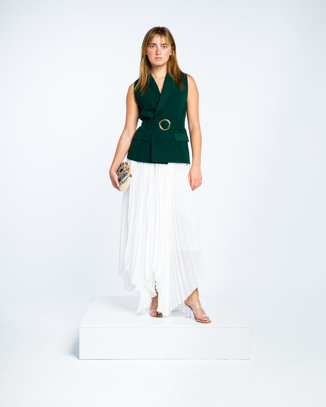 Woman wearing dark green sleeveless belted blazer and white pleated asymmetrical skirt