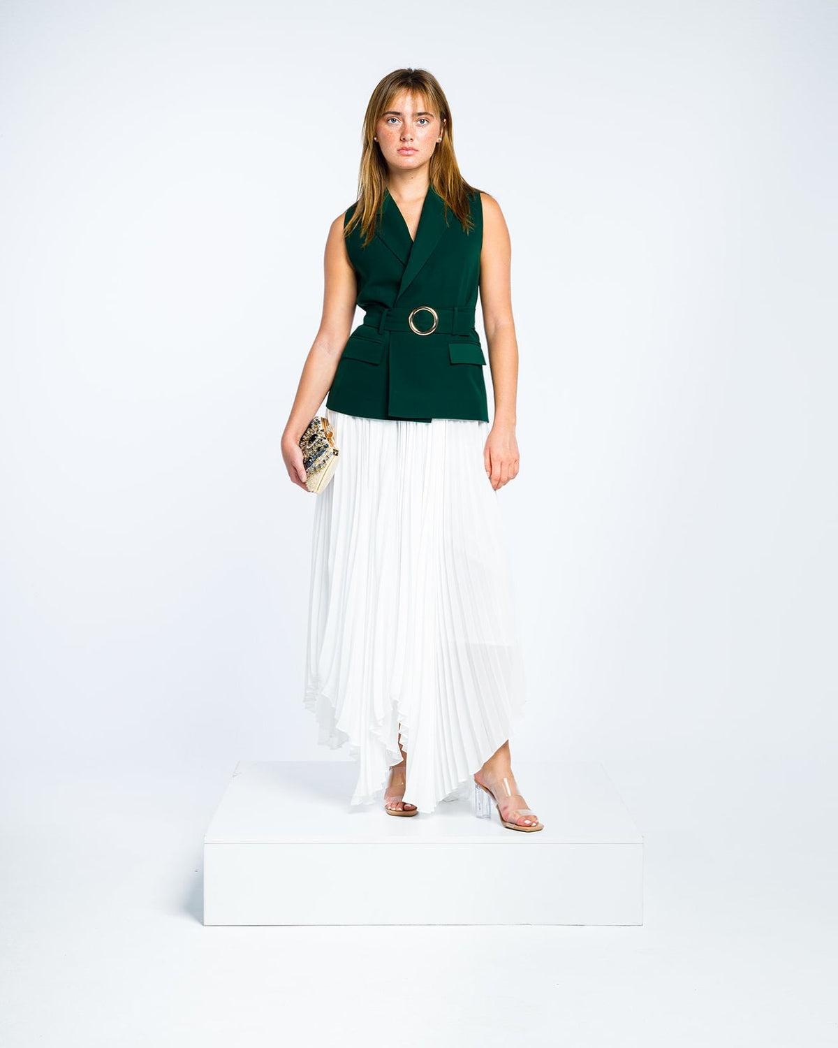 Woman wearing dark green sleeveless belted blazer and white pleated asymmetrical skirt