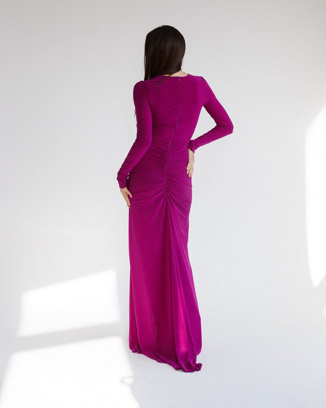 Woman in a vibrant purple long-sleeve evening gown with ruched detailing, standing indoors