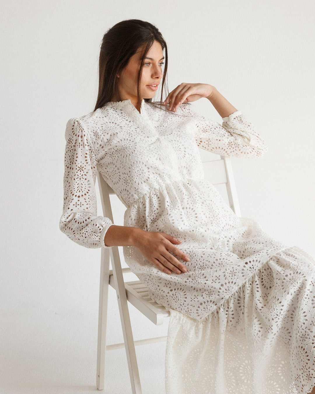 Woman in a white lace long-sleeve maxi dress with broderie anglaise details, sitting on a chair