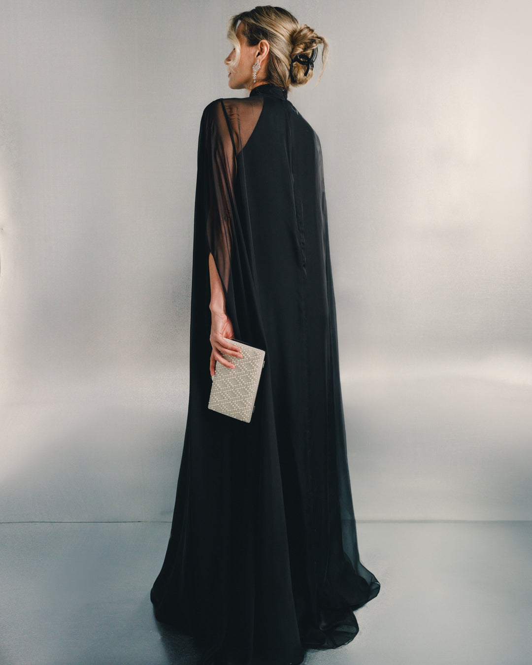 Woman in elegant black sheer evening gown holding a pearl clutch against a silver background