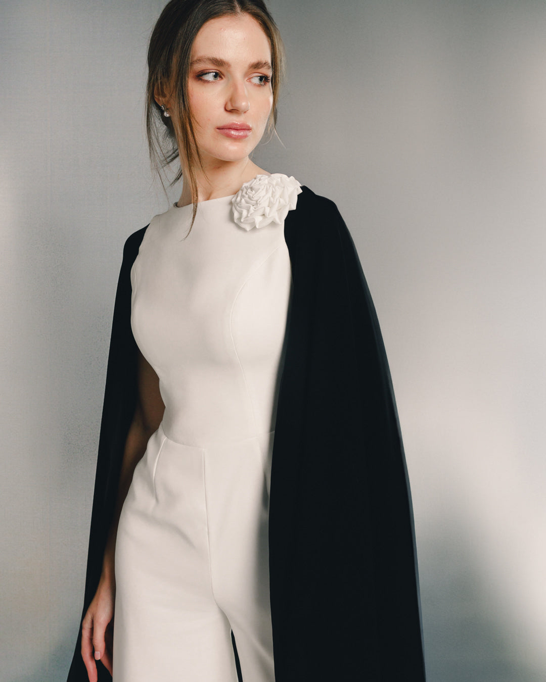 Woman in elegant white jumpsuit with floral shoulder detail and black cape