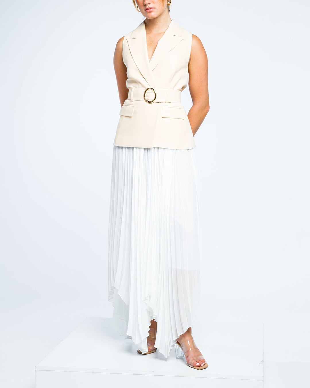 Woman in sleeveless beige belted blazer and white pleated maxi skirt on white background